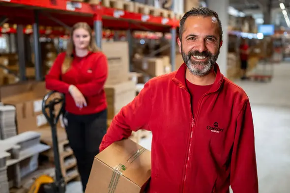 Um homem com barba, sorrindo e vestindo uma camiseta vermelha, segura uma caixa em um armazém. Ao fundo, uma mulher também em camiseta vermelha está posicionada ao lado de um carrinho de transpporte. O ambiente possui prateleiras cheias de caixas.