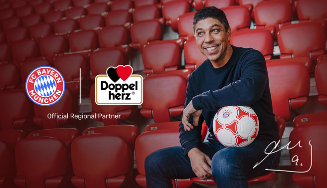 Homem sorridente sentado em um estádio vazio, segurando uma bola de futebol. Ao fundo, logos do FC Bayern München e Doppelherz, com a inscrição "Official Regional Partner". O homem usa um suéter escuro e parece estar em um ambiente de apoio ao futebol.