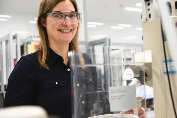 Mulher sorridente usando óculos de proteção, em um ambiente de laboratório. Ela está próxima a um equipamento, com um fundo claro e diversos equipamentos de laboratório visíveis ao fundo.