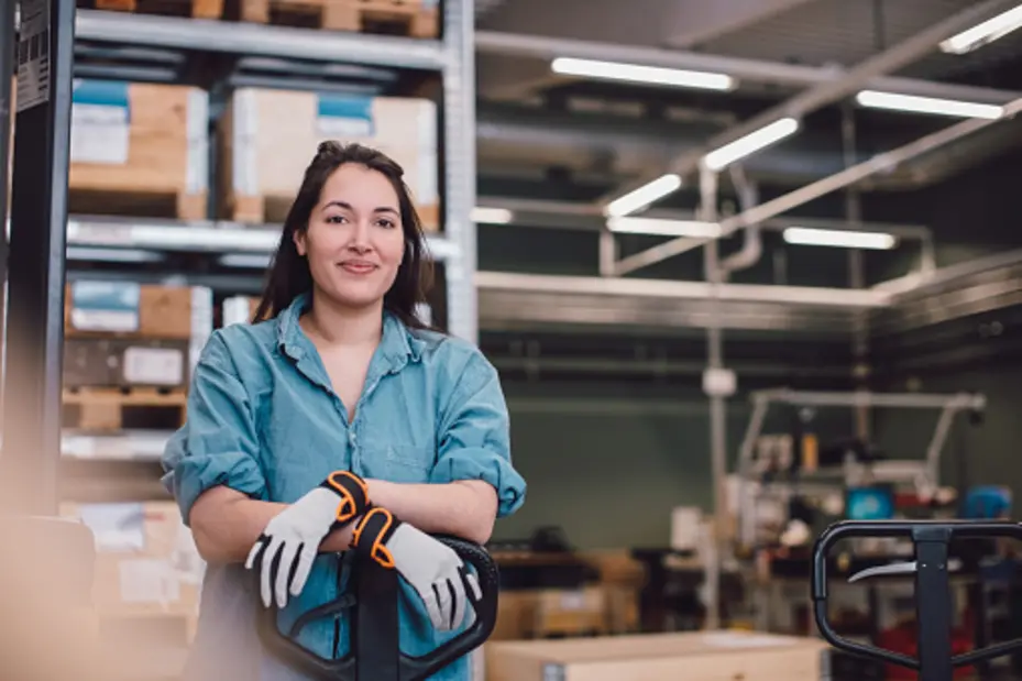 Mulher com luvas de trabalho sorri enquanto se apoia em um carrinho de transporte em um ambiente industrial. Ao fundo, prateleiras com caixas e equipamentos de trabalho.