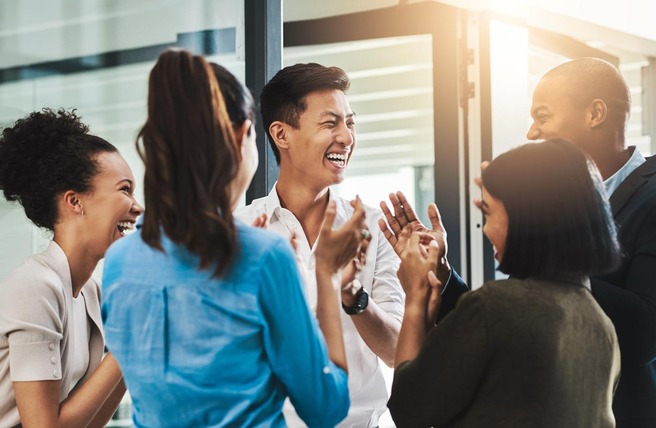 Grupo diversificado de seis pessoas, sorrindo e se divertindo, enquanto conversam animadamente em um ambiente interno. O clima é de celebração e alegria, com palmas e risos. A luz natural entra pelos janelões ao fundo.