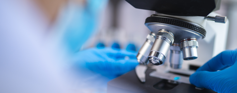 Imagem de um microscópio sendo manuseado por uma pessoa com luvas azuis. O foco está nas lentes do microscópio e na mão que ajustar o equipamento. O fundo é desfocado, sugerindo um ambiente de laboratório.