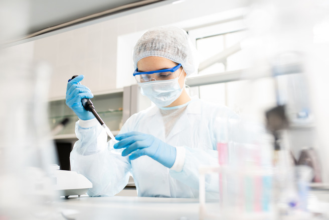Uma profissional de laboratório, usando avental, máscara e luvas, está manipulando uma pipeta em um ambiente de pesquisa. O foco está na tarefa que ela realiza, com equipamentos de laboratório visíveis ao fundo.