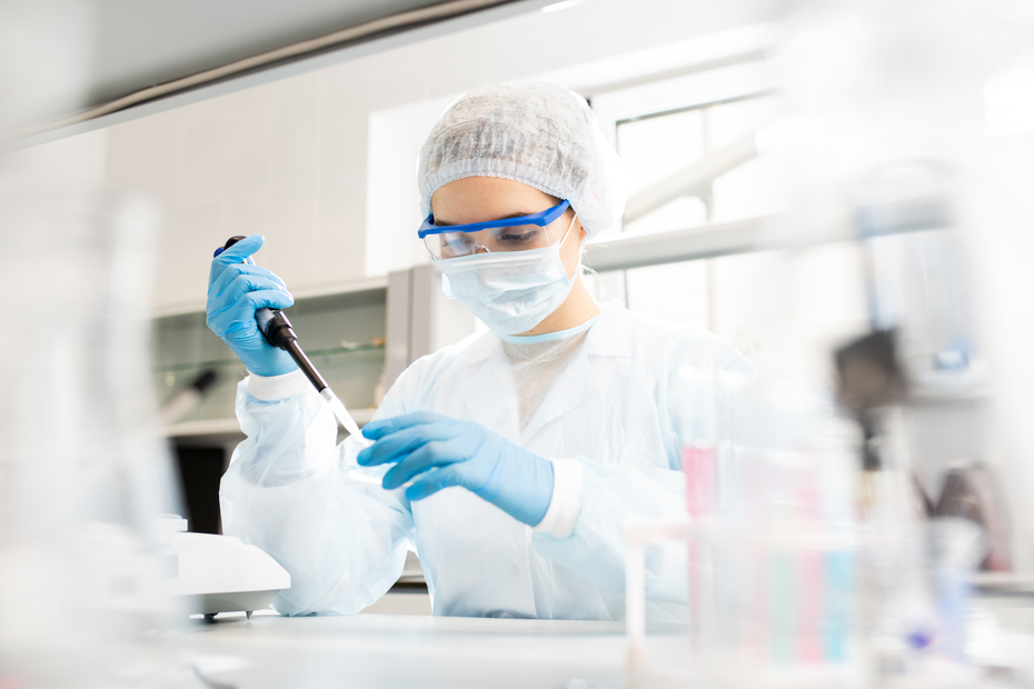 Uma profissional de laboratório, usando avental, máscara e luvas, está manipulando uma pipeta em um ambiente de pesquisa. O foco está na tarefa que ela realiza, com equipamentos de laboratório visíveis ao fundo.