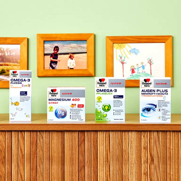 Em uma prateleira de madeira, são exibidos quatro produtos de saúde da marca Doppelherz: Omega-3, Magnesium 400 Citrato, Omega-3 Pluranzlich e Augen Plus. Ao fundo, há fotos de crianças e desenhos emoldurados em uma parede verde.