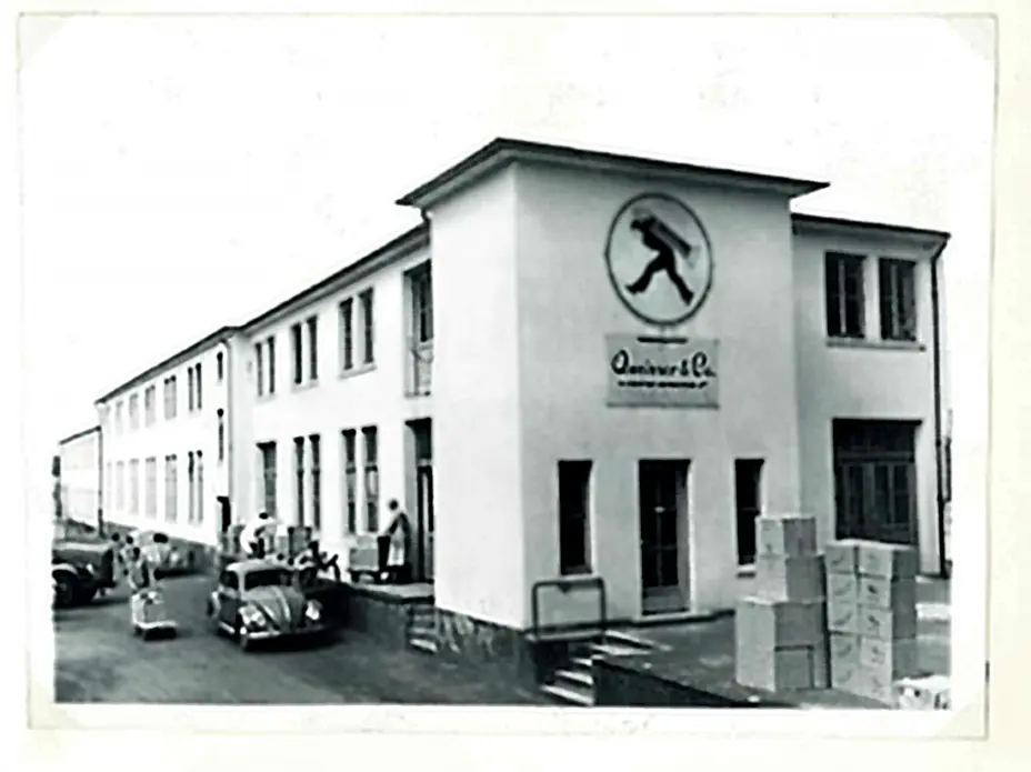 Fachada de um edifício histórico, com uma logo preta de um homem caminhando. A construção é de dois andares, com várias janelas e portas. Há caixas empilhadas na frente e alguns carros antigos estacionados. A imagem é em preto e branco.