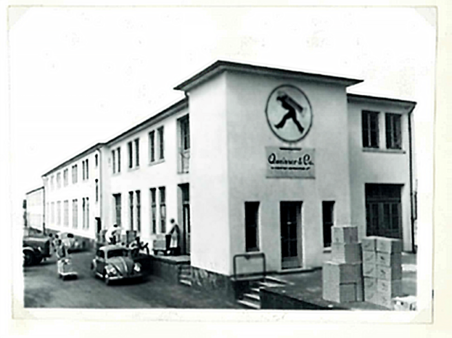 Fachada de um edifício histórico, com uma logo preta de um homem caminhando. A construção é de dois andares, com várias janelas e portas. Há caixas empilhadas na frente e alguns carros antigos estacionados. A imagem é em preto e branco.