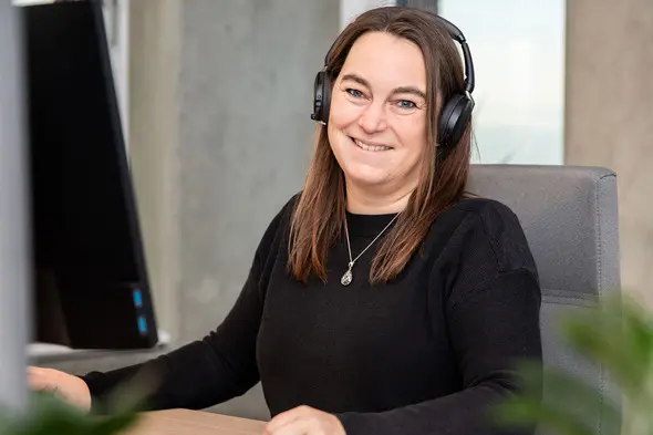 Mulher sorridente usando fones de ouvido em um ambiente de escritório. Ela está sentada diante de um computador e tem cabelos castanhos. A sala é iluminada e moderna, com elementos de plantas ao fundo.