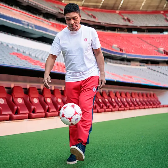 Homem vestido com camiseta branca e calças vermelhas pratica dribles com uma bola de futebol em um estádio vazio. Ele está sorrindo e caminhando ao lado de um banco de reservas. O campo é bem iluminado, evidenciando a atmosfera esportiva.