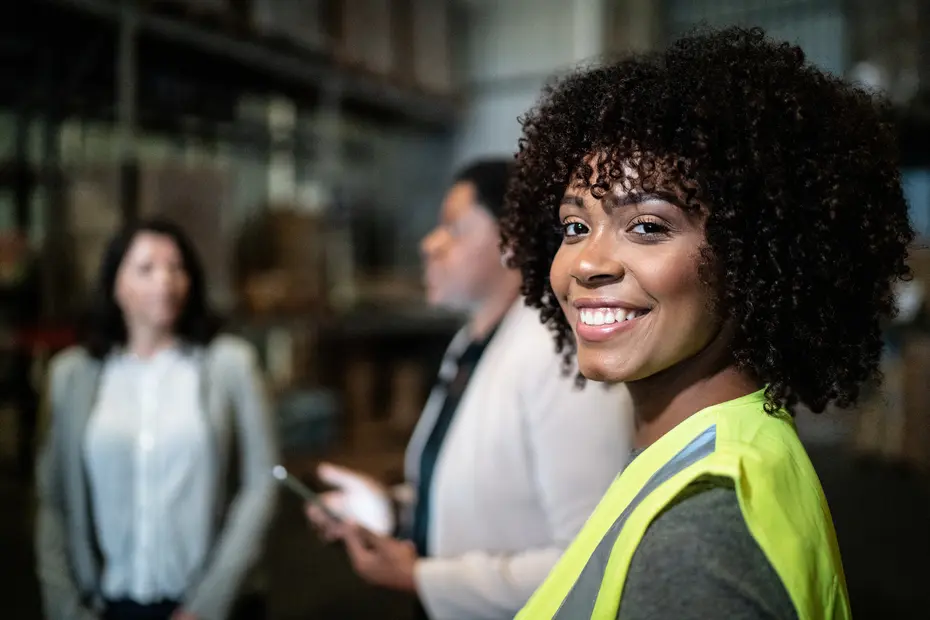 Uma mulher sorridente com cabelos cacheados usa um colete amarelo e está em um ambiente de armazém. Ela está olhando para a câmera, enquanto duas outras mulheres conversam ao fundo. A cena transmite um ambiente de trabalho colaborativo.