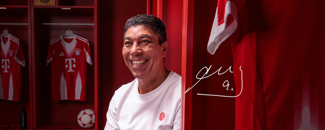Um homem sorridente está sentado em um vestiário, com camisas do Bayern de Munique visíveis em armários vermelhos ao fundo. Ele veste uma camiseta branca e há uma bola de futebol ao seu lado. A imagem tem um ambiente acolhedor e esportivo.