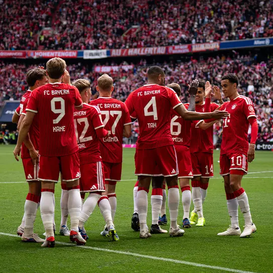 Jogadores do FC Bayern se reúnem em campo para comemorar, vestidos com uniformes vermelhos e brancos. Eles estão em posição de conversa, sorrindo e se dando boas-vindas, enquanto a torcida está ao fundo, criando uma atmosfera animada.