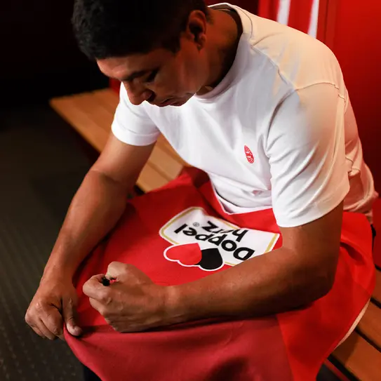 Homem sentado em um vestiário, vestindo uma camiseta branca e preparando um tecido vermelho. Ele parece concentrado, segurando o tecido com uma mão e ajustando a estampagem que diz "Pogger Fk". O ambiente é simples, com painéis vermelhos ao fundo.