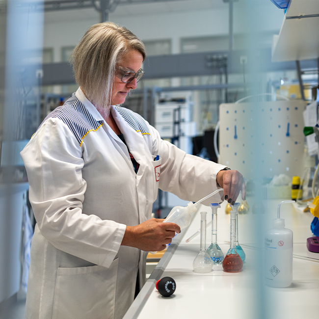 Uma pesquisadora de cabelo grisalho, usando um jaleco branco, está em um laboratório. Ela está segurando uma pipeta e concentrada em transferir líquido de um frasco para outro, com equipamentos de laboratório ao fundo.