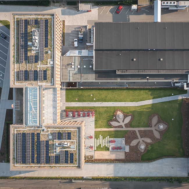Vista aérea de um complexo arquitetônico com telhados cobertos de painéis solares. No chão, um jardim com caminhos sinuosos e áreas de lazer, rodeado por edifícios modernos e áreas de estacionamento.