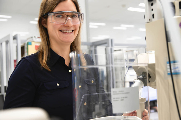 Mulher sorridente usando óculos de proteção, em um ambiente de laboratório. Ela está próxima a um equipamento, com um fundo claro e diversos equipamentos de laboratório visíveis ao fundo.